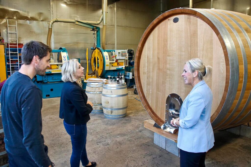 Zwei Frauen und ein Mann stehen in einer Werkstatt vor einem großen Holzfass und unterhalten sich.