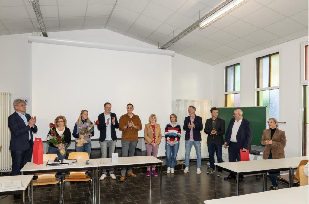 Eine Gruppe von Personen steht in einem Seminarraum und applaudiert. Zwei Frauen halten Blumensträuße in der Hand.