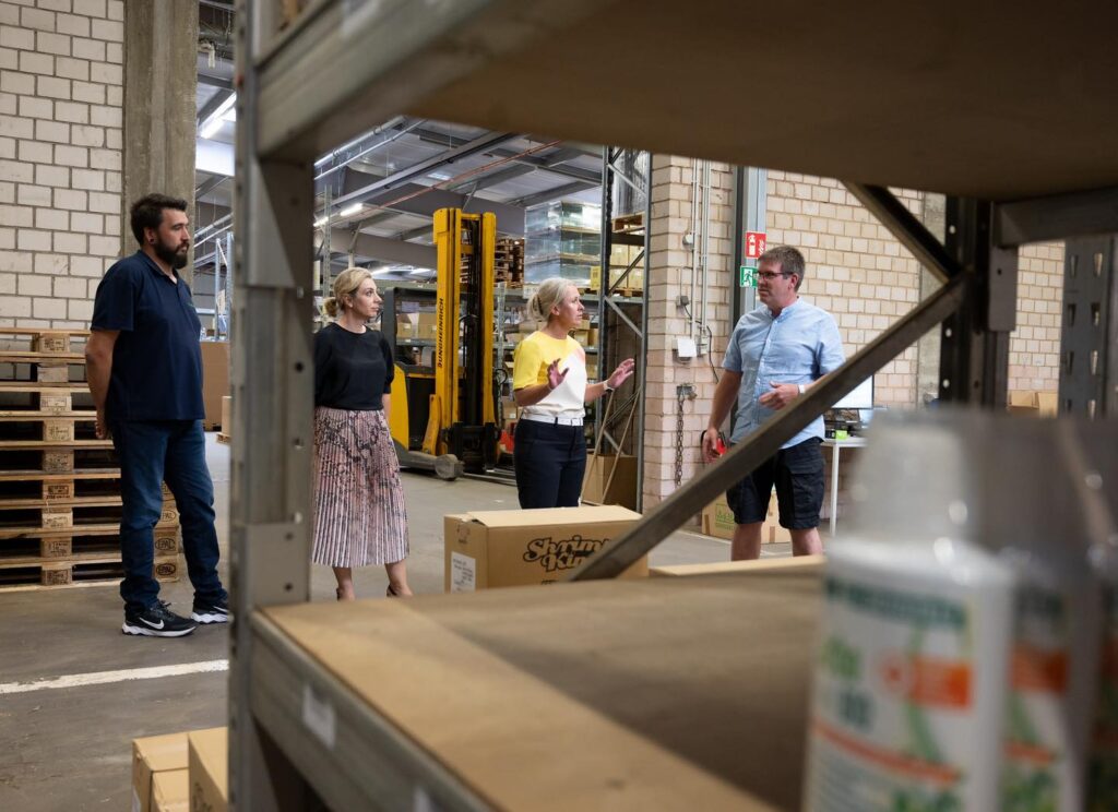 Vier Personen stehen in einem Lager und diskutieren. Im Hintergrund sind Regale, Kartons und ein Gabelstapler zu sehen.