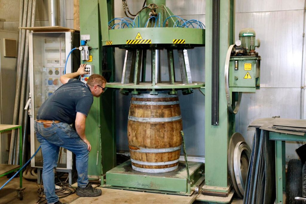 Ein Handwerker kontrolliert ein Holzfass in einer großen hydraulischen Presse in einer Werkstatt.