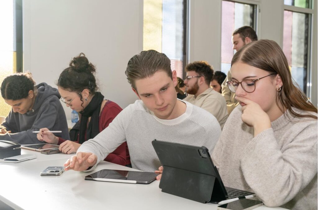 Zwei Schüler sitzen mit einem Tablet und arbeiten konzentriert, während andere im Hintergrund zu sehen sind.