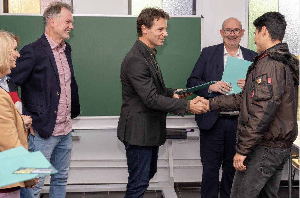 Ein Mann überreicht einem Teilnehmer ein Zertifikat und schüttelt ihm die Hand. Weitere Personen stehen im Hintergrund.